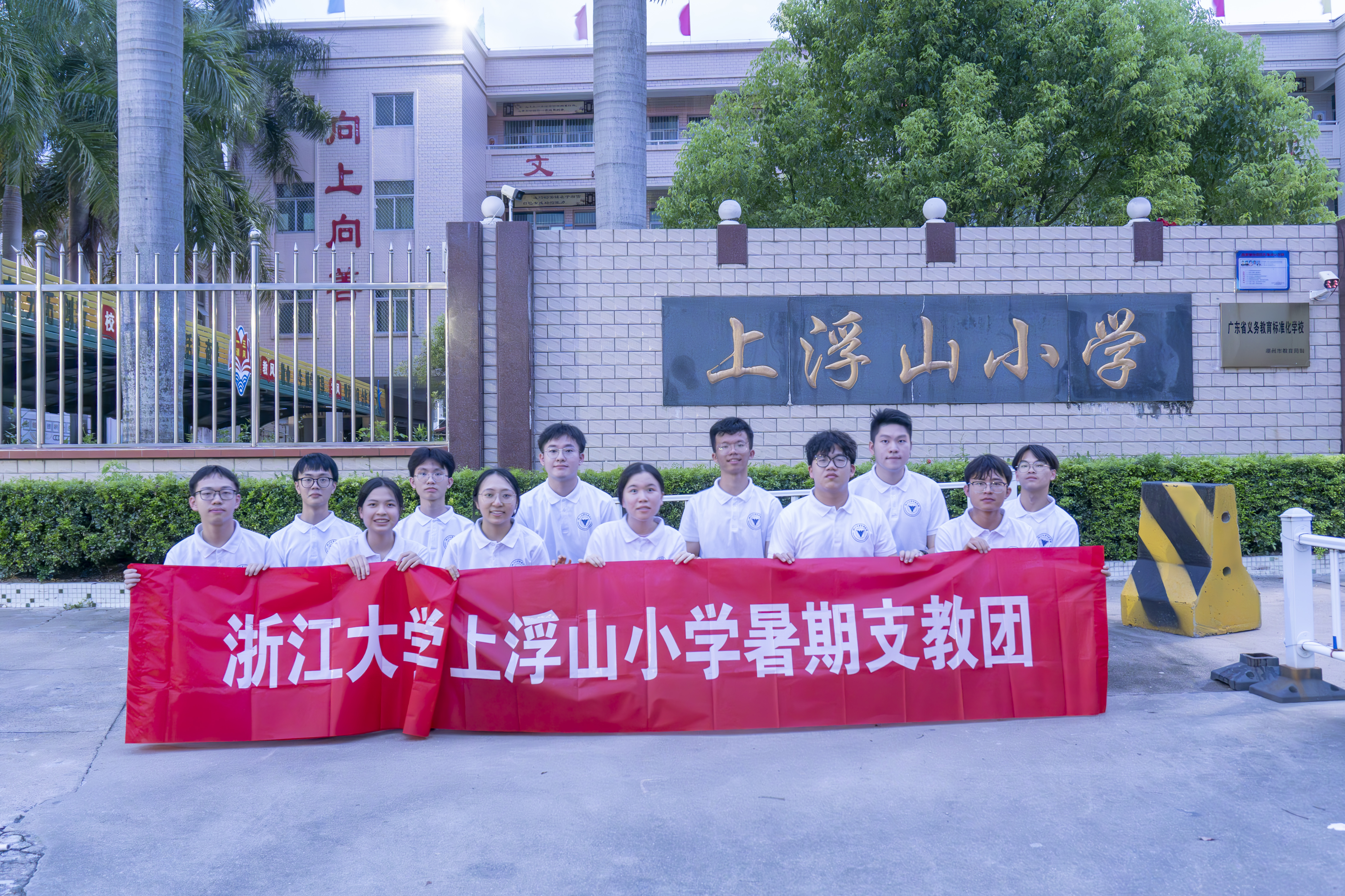 浙江大学上浮山小学暑期支教团成员身穿白色队服，手持红色横幅合影，横幅文字为：浙江大学上浮山小学暑期支教团。背景为上浮山小学正门，墙上有金色校名，上方有绿树和教学楼，环境充满夏日气息，氛围温馨而充满团队凝聚力。